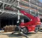 Front view of red Magni telehandler with an extended boom and stabilizers on a construction site in front of steel framework.