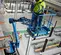 Top angled view of an operator in safety gear standing on a narrow blue boom lift platform working on a construction site.