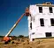 Side view of an orange boom lift with operator working on a two-story house under construction, on uneven dirt terrain.
