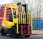 A man operates a yellow Hyster H50 forklift with red forks, positioned on a paved surface, with tall stacks of red Coca-Cola crates to the left and blue water bottle crates to the right, under an overcast sky. The forklift is viewed from the front-right side.