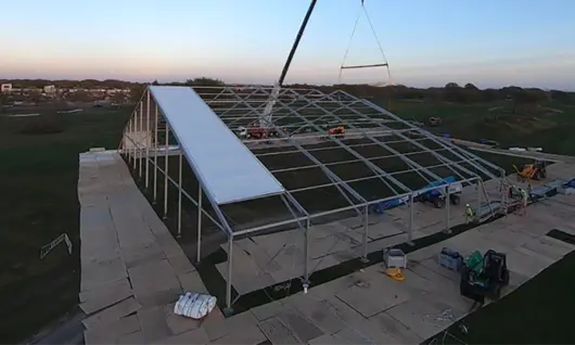 Una grúa instalando un panel de techo en una gran estructura de tela temporal al atardecer en un campo abierto