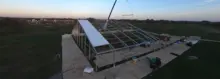 Una grúa instalando un panel de techo en una gran estructura de tela temporal al atardecer en un campo abierto